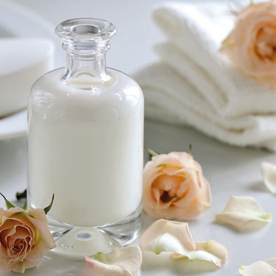 Eine elegante Glasflasche mit weißer Lotion steht auf einem hellen Untergrund. Umgeben ist sie von zarten Rosenblüten und weißen Handtüchern, was eine ruhige, pflegende Spa-Atmosphäre vermittelt.