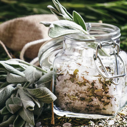 Ein Glas mit aromatisiertem Badesalz, gefüllt mit Kräutern und Meersalz, steht auf einem Holztisch, umgeben von frischen Salbeiblättern und getrockneten Pflanzenteilen.