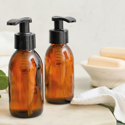 Zwei braune Spenderflaschen mit schwarzem Pumpverschluss stehen auf einem weißen Untergrund, daneben Handtücher, Eukalyptusblätter und eine Seifenschale.