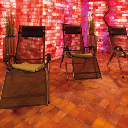 Mehrere Relaxliegen stehen in einem stimmungsvoll beleuchteten Ruheraum mit roten Salzwänden und warmem Licht. Der Boden ist mit Salzsteinen ausgelegt, an den Wänden stehen dekorative Pflanzen.