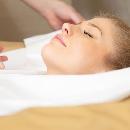 Eine entspannte Frau liegt mit geschlossenen Augen auf einer Behandlungsliege, eingehüllt in ein weißes Tuch, während eine Hand sanft ihren Kopf oder Nackenbereich berührt.