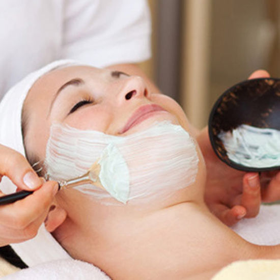 Eine Frau liegt entspannt mit geschlossenem Auge, während ihr eine kosmetische Gesichtsmaske mit einem Pinsel aufgetragen wird. Sie lächelt leicht und trägt ein weißes Haarband.