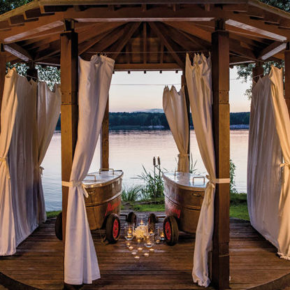 Romantischer Pavillon mit weißen Vorhängen und zwei nostalgischen Badewannen auf Holzplattform, beleuchtet von Kerzenlicht mit Blick auf einen ruhigen See bei Sonnenuntergang.