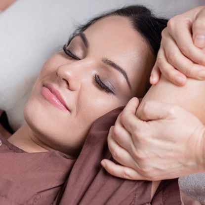 Zufriedene Frau mit geschlossenen Augen in brauner Wellnesskleidung genießt eine entspannende Armmassage in ruhiger Wohlfühlatmosphäre.