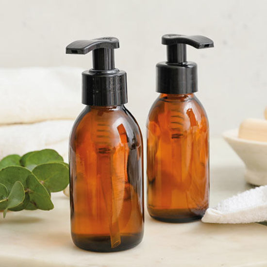Zwei braune Spenderflaschen mit schwarzem Pumpverschluss stehen auf einem weißen Untergrund, daneben Handtücher, Eukalyptusblätter und eine Seifenschale.