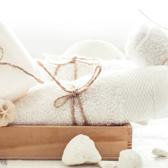 Weißes Handtuchset mit dekorativer Kordel auf einem Holztablett, daneben weiße Naturseife und Bimssteine.