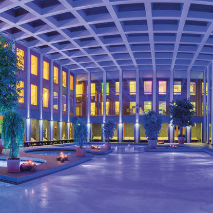 Abendlicher Innenbereich der SaarowTherme mit beleuchtetem Solebecken. Lila gefärbtes Wasser durch Unterwasserscheinwerfer, dekoriert mit Grünpflanzen und brennenden Kerzen am Beckenrand. Goldenes Licht fällt durch die Glasfront im oberen Bereich des Gebäudes.