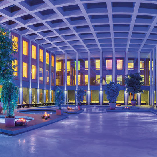 Abendlicher Innenbereich der SaarowTherme mit beleuchtetem Solebecken. Lila gefärbtes Wasser durch Unterwasserscheinwerfer, dekoriert mit Grünpflanzen und brennenden Kerzen am Beckenrand. Goldenes Licht fällt durch die Glasfront im oberen Bereich des Gebäudes.