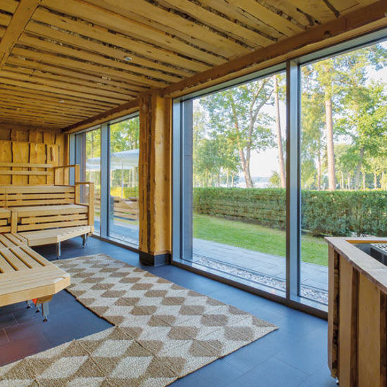 Große, moderne BrotBackSauna mit Holzverkleidung und mehreren Sitzebenen; durch die Panoramafenster blickt man auf eine grüne Außenanlage mit Büschen und Bäumen.