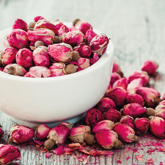 Eine weiße Schale gefüllt mit getrockneten Rosenknospen in kräftigem Pink, umgeben von weiteren Rosenknospen und Blütenblättern auf einem rustikalen Holztisch.