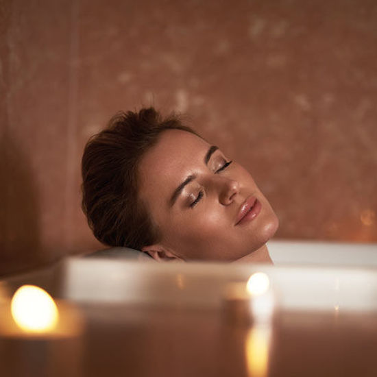 Eine Frau entspannt in einer Badewanne bei Kerzenschein, lehnt mit geschlossenen Augen den Kopf an den Rand.